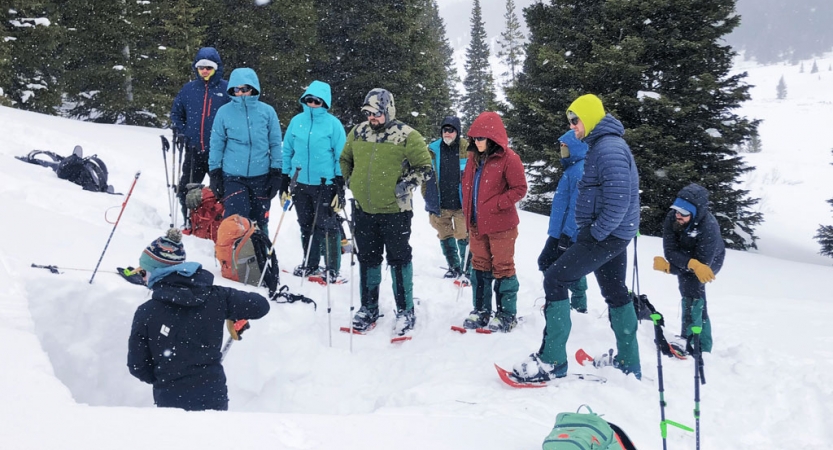 A group of people wearing winter gear watch an instructor practice avalanche training in snow. 